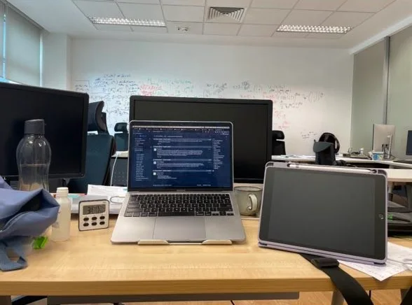 My desk in the group’s office at the Centre for Quantum Technologies: a laptop on a stand, an iPad, a water bottle and a kitchen timer, with a whiteboard wall of equations behind.