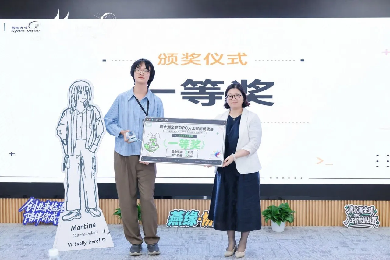 The award ceremony: Siyu Wang holding the first-prize board with a presenter, beside a life-size cardboard cutout of Martina labelled “Co-founder, virtually here!”.